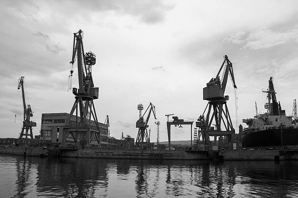 Black and white view of a harbor showcasing industrial infrastructure, including towering cargo cranes and a docked ship, against a cloudy sky reflecting on calm waters