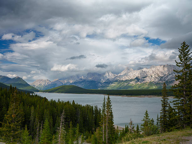 Stunning view of a pristine lake surrounded by evergreen forest and towering mountains in Peter Lougheed Provincial Park, Alberta, Canada. The dramatic clouds and sunlight playing on the rugged peaks create a powerful contrast, showcasing the raw beauty of the Canadian Rockies. Perfect for themes related to outdoor travel, untouched nature, Canadian parks, hiking adventures, and alpine scenery.