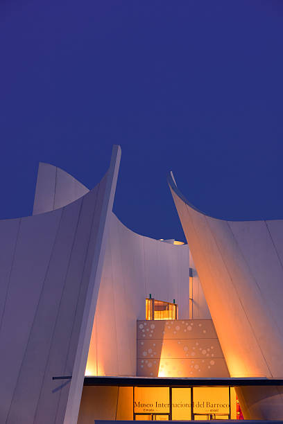 Puebla, Pue., Mexico - March 27, 2021: shot in the evening with rays from passing cars on the walls of entrance Museo Internacional del Barroco in the evening. the walls are illuminated with yellow lamps. the sky is dark - the road is concrete, the walls are made of tiles.