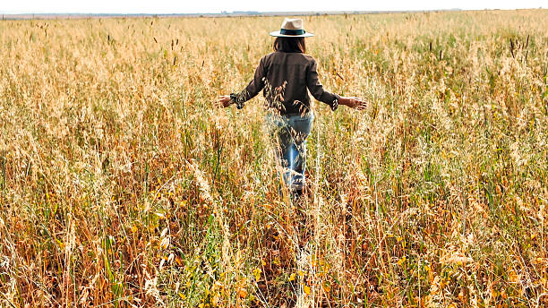 A woman in her 30s walks alone through a rural agricultural field filled with tall grasses or grains, possibly oats or wheat. She wears a wide-brimmed hat, casual clothing, and has light skin, conveying a connection to nature and rural life. The photo was taken during the daytime under a bright but cloudy sky, indicating overcast weather. The scene reflects simplicity, countryside lifestyle, and sustainable farming.