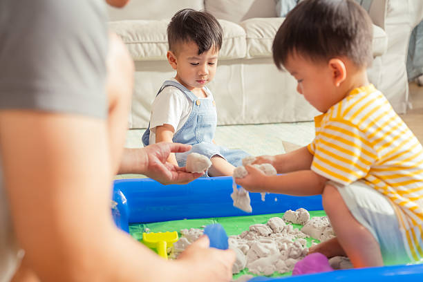 istock Two toddler boys are deeply engaged in playing with hydrophobic sand inside a clear plastic container, set on the floor in a sunlit living room. One boy gently sifts the sand through his fingers while the other molds it with curiosity. The moment 2222909178