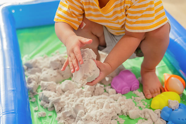 Two toddler boys are deeply engaged in playing with hydrophobic sand inside a clear plastic container, set on the floor in a sunlit living room. One boy gently sifts the sand through his fingers while the other molds it with curiosity. The moment highlights early STEM learning through tactile exploration and imaginative play. Surrounded by toys and home comforts, this indoor activity sparks creativity, fine motor skills, and childhood joy in a safe, nurturing environment.