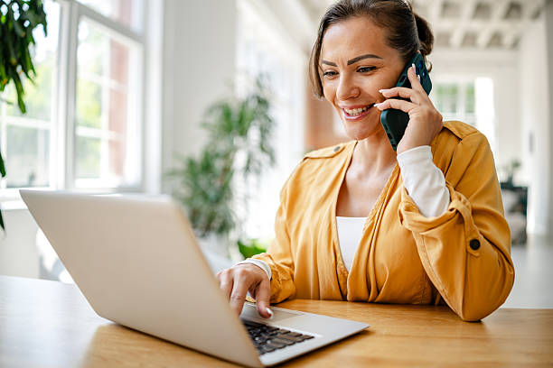 istock Woman in yellow shirt talks on phone while working on laptop in a bright office space 2222913207