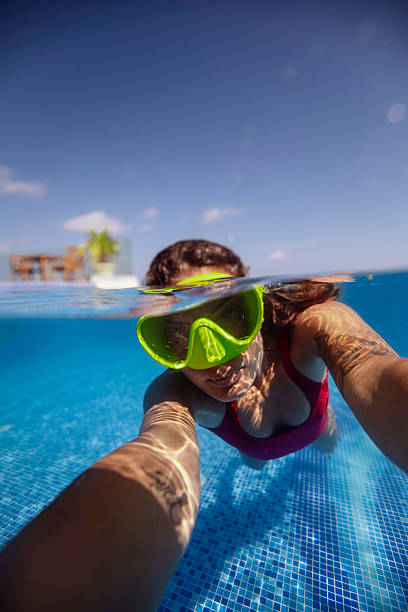 A woman wearing swim goggles and a red swimsuit snorkels in a clear turquoise swimming pool under a sunny blue sky and taking selfie