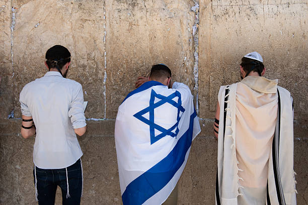Three Jewish men - one wrapped in an Israeli flag, one wearing a prayer shawl or tallit and one with only a white shirt, pray at the Western Wall in Jerusalem on Israeli Independence Day. stock photo