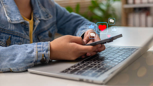 Using a smartphone while a laptop is open, with social media icons floating, such as likes, hearts, and comments, representing online interaction and digital lifestyle.