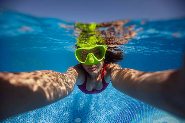 A woman in a swimsuit and goggles swims underwater in a clear blue pool on a sunny day, capturing a fun summer atmosphere