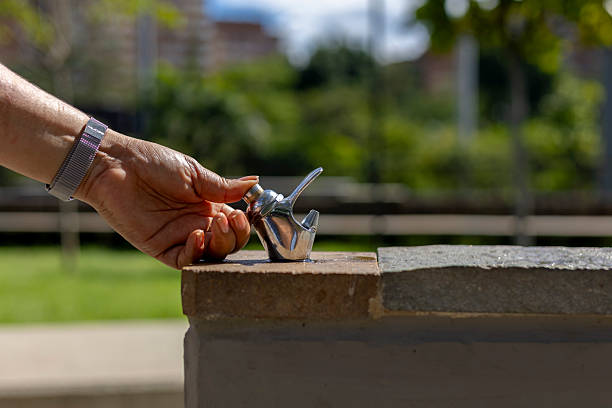 A close-up image of a person's hand pressing the button of a public drinking fountain, with a clear stream of water arching into the air. The background shows a lush, green park setting, highlighting themes of hydration, public amenities, outdoor health, and environmental awareness.