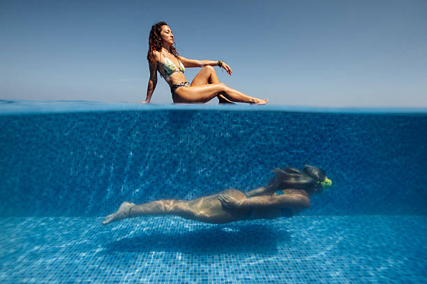 A serene poolside moment with a woman sunbathing on the edge while another woman swims underwater in clear blue waters