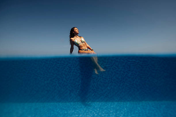 Attractive young woman in bikini leaning on the edge of infinity swimming pool. Beautiful female in a swimming pool looking away on a summer day.