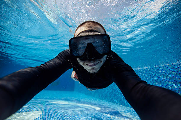 A man, wearing a black diving suit and goggles, smiles while swimming underwater in a clear blue pool, surrounded by shimmering tile patterns