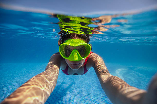 Underwater view of a woman wearing swim goggles in a clear blue swimming pool on a sunny day and taking selfie