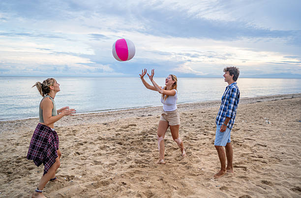 Three young adults playing beach volleyball on a sandy shore, laughing and enjoying their time together under a cloudy sky