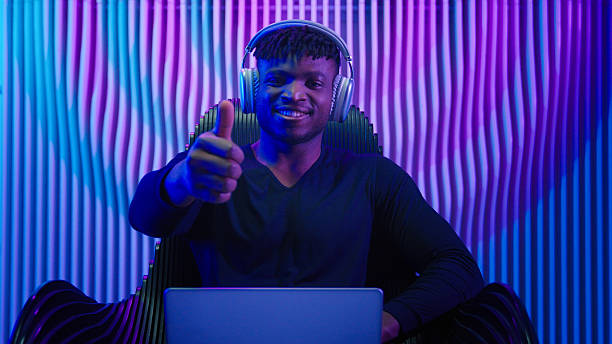 African American man guy male coder gamer programmer in chair wear headphones using laptop online work crypto software play internet game modern technology neon futurism showing thumb up like gesture. High quality 4k footage