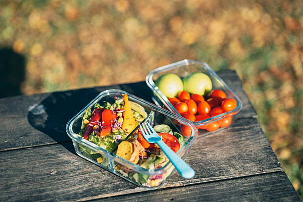 glass containers rest on a wooden bench outdoors. The foreground container holds a vibrant salad with lettuce, bell peppers, and shredded carrots; beside it, another box contains cherry tomatoes and green apples. Warm sunlight filters onto the lawn, creating a relaxed, natural vibe. This image conveys how modern individuals embrace homemade organic light meals to practice healthy eating, minimize single-use plastics, and pursue sustainable living.