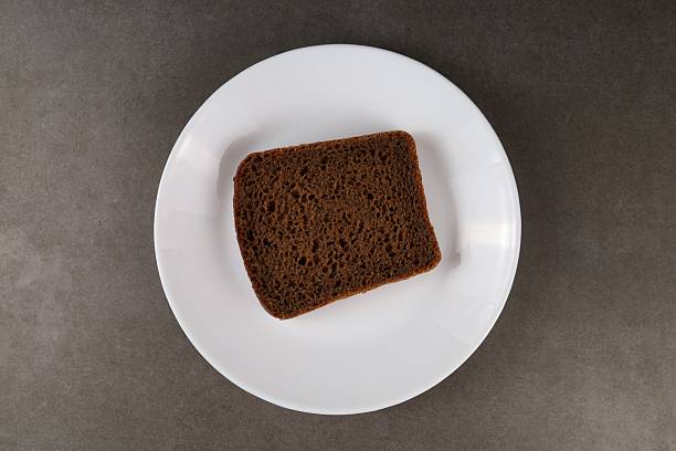 A slice of dark bread is placed on a plain white plate against a dark surface, showcasing its texture and color.