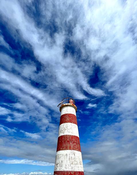 Red and white lighthouse rising against a vibrant blue sky filled with dynamic white clouds, highlighting the contrast between the structure and the Faro seascape