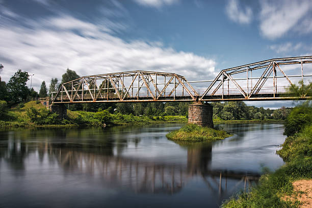 A striking view of the historic iron bridge in Valmiera, Latvia, spanning the calm waters of the Gauja River. Captured under a dynamic sky with long-exposure effect, the image highlights the bridge's rustic steel structure and its reflection in the water. A perfect representation of Latvian heritage, scenic architecture, and tranquil nature—ideal for travel, infrastructure, or European landscape themes.