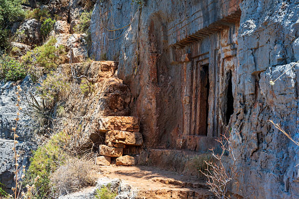 An ancient Lycian rock tomb carved into a steep cliff face, accessed by rough-hewn stone steps nestled among Mediterranean shrubs. The weathered reliefs around the tomb entrance reflect the architectural finesse of the Lycian civilization, set against the dramatic textures of the rugged mountain terrain.