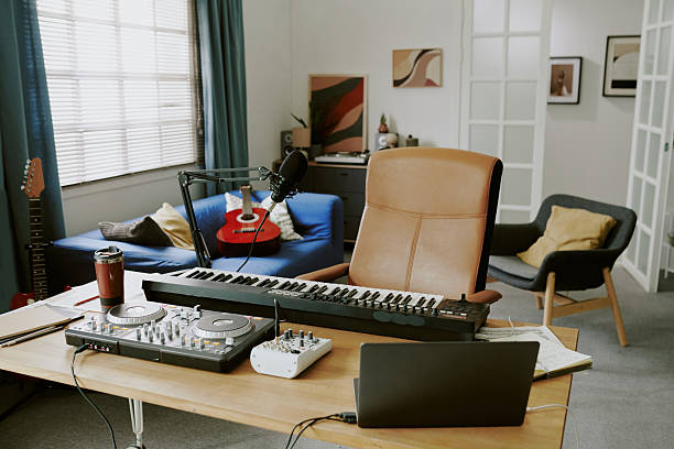 Modern home studio showing digital piano, DJ controller, audio mixer, microphone, laptop on desk with acoustic guitar on sofa in background, creative workspace for music recording and production