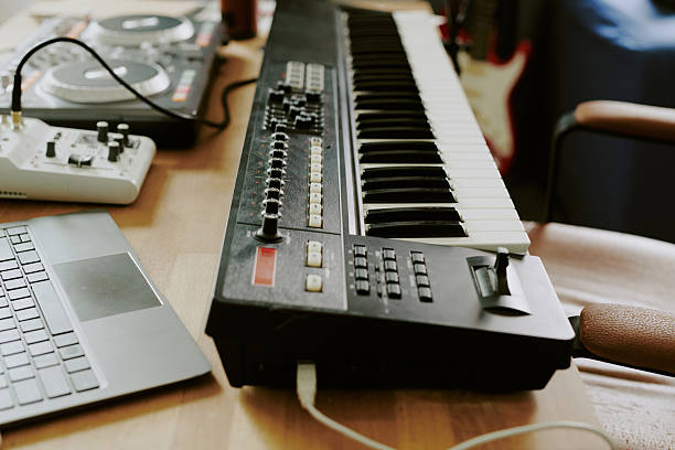 Electronic music production equipment including digital keyboard synthesizer, DJ controller, audio interface and laptop arranged on wooden desk in home studio environment