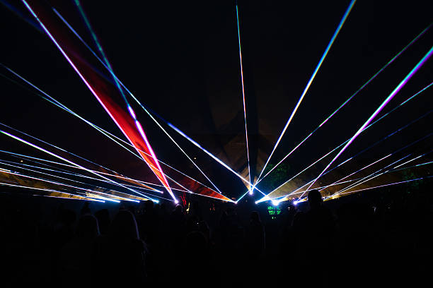 Vibrant laser beams form dynamic patterns in the dark night, with silhouetted people in the foreground and faint structures or trees in the distance.