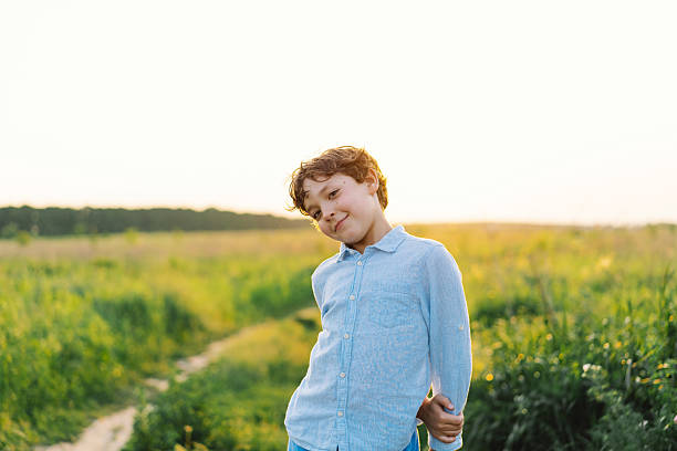 A joyful child stands in a vibrant green field during sunset, smiling and playfully posing with a relaxed demeanor. The warm glow highlights the serene landscape.