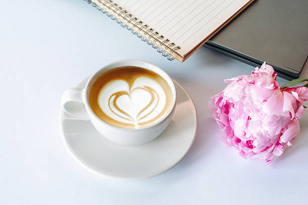 A cup of cappuccino, notebook and peony flower on white background. Top view.