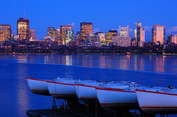 Small sailboats are moored along an icy frozen Charles River while the lights of the Boston skyline is reflected at night