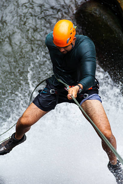 A climber (not recognizable, silhouette only) is rappelling / abseiling down a cliff in front of water and mountains at sunset. The rope silhouette is also visible.
