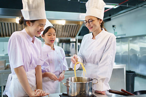 Two female students in chef uniforms attentively listen to a cooking teacher in a large kitchen. The instructor demonstrates how to boil spaghetti noodles for a culinary class activity.