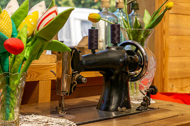 Vintage sewing machine placed on a wooden table, surrounded by decorative fabric and glass tulips. Rustic interior in the background with wooden elements and bottles used as vases. Nostalgic, handmade, and vintage atmosphere.