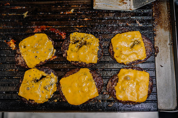 Liepa, Latvia - June 27, 2025 - Six beef burger patties with melted cheddar cheese cooking on a greasy grill, viewed from above in a food preparation setting.