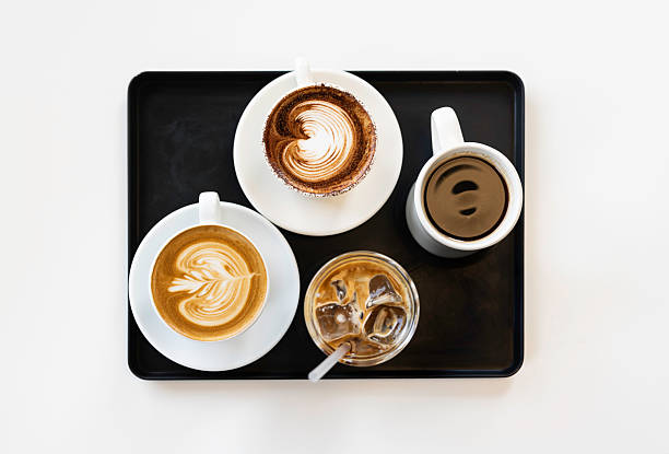 A top-down view of four different coffee drinks — hot latte with latte art, cappuccino, iced latte, and black coffee — served in white cups and glass on a minimalist black tray over a white table. Perfect for use in content related to cafes, baristas, beverages, and modern food styling.