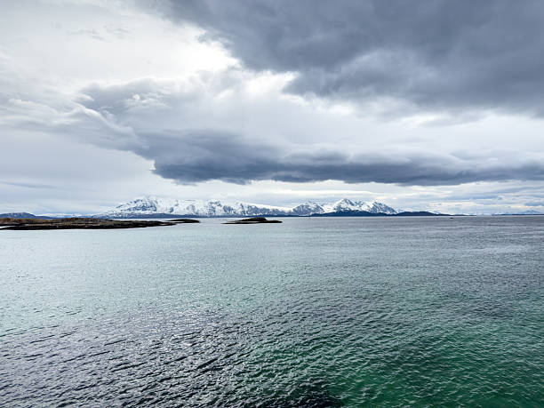 Tranquil ocean water with small rocky island and lighthouse under cloudy sky. Peaceful coastal landscape with distant mountains on horizon. Serene sea view for nature and travel themes.