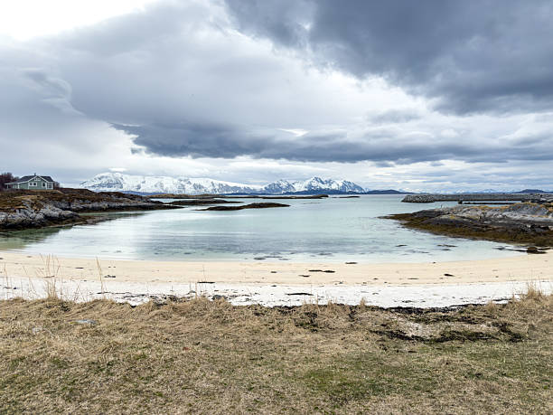 Calm coastal bay with clear water and sandy beach under cloudy sky. Snow-covered mountains in the distance frame the peaceful shoreline. Scenic nature landscape with rocky coast and isolated house.