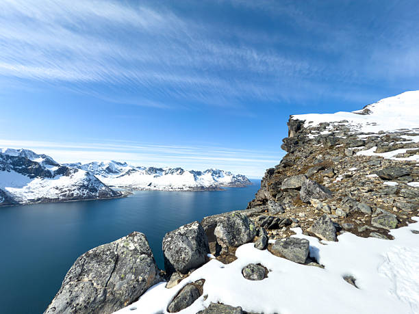 Snow-covered rocky cliff with large boulders overlooking calm blue ocean and distant snow-capped mountains. Scenic winter landscape with clear sky and rugged natural terrain. Mountain and coastal nature view.
