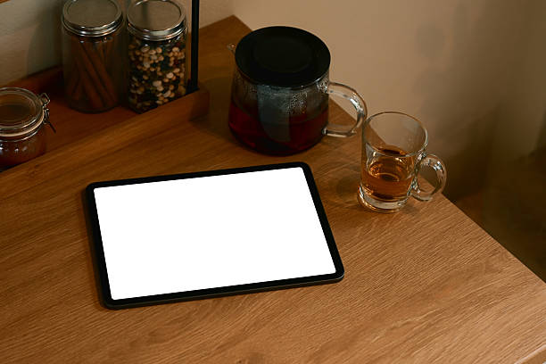 Digital tablet with blank screen on kitchen table beside glass teapot and cup of tea.