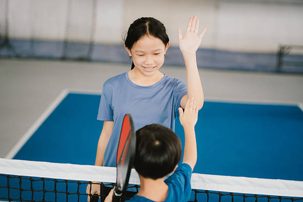 Two children, a girl and a boy, are seen giving each other a high-five across a tennis net, celebrating a fun and friendly match. The girl is smiling and raising her hand confidently, while the boy, holding a tennis racket, reaches up to meet her gesture.