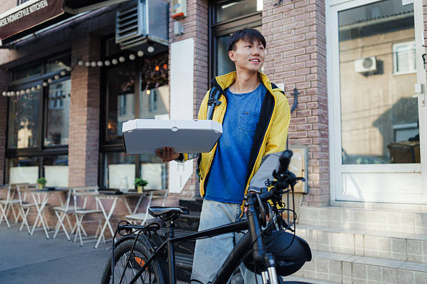 A smiling young Chinese delivery man holding a food delivery box, standing beside a bicycle outdoors.