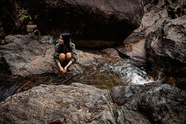 A young woman exploring the forest of a national park in Koh Phangan, Thailand. She rests by the spring, her gaze directed toward the horizon, wearing a quiet smile that shows her deep appreciation for the natural beauty surrounding her.