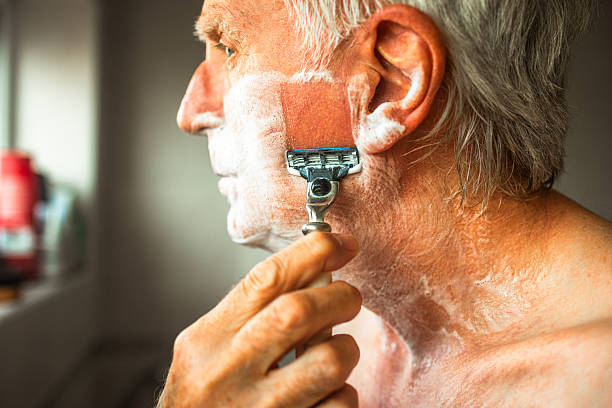 Close up portrait depicting a senior man in his 70s wet shaving his face with a razor. His face has a lather of shaving cream on it to ease the process.
