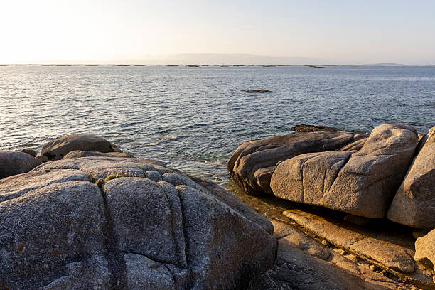 A rocky shoreline with a large rock in the foreground. The water is calm and the sky is a light blue