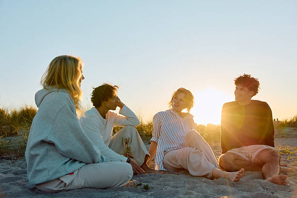 istock Four cheerful friends sitting on beach talking at sunset, backlit 2223044582