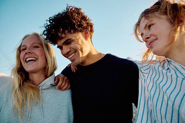 Three cheerful young adult friends smiling with arms around each other, low angle stock photo