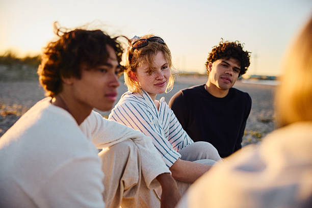 istock Group of young people sitting and talking on a sandy lakeside beach at sunset 2223045169