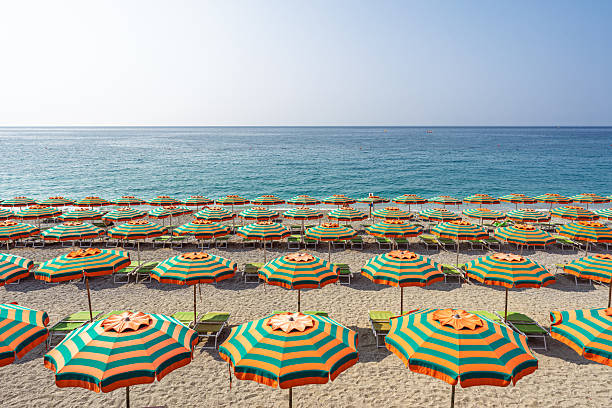 Vibrant orange and green umbrellas line the sandy shore on a clear morning along the Ligurian coast in Italy, facing the calm turquoise sea