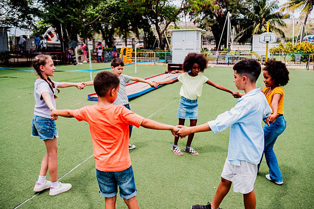 Group of Caucasian children walking on grass with female school teacher supervising in park, kids participating in outdoor activity together surrounded by green trees
