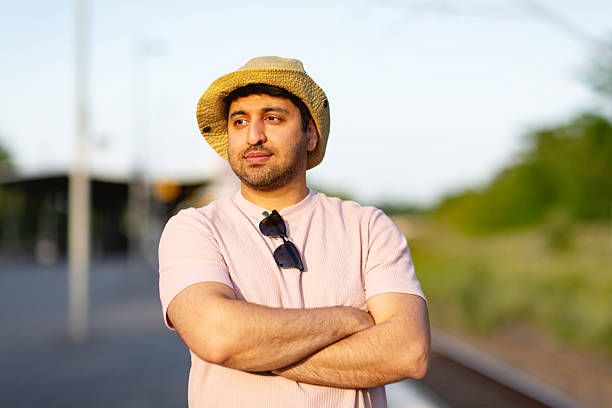 Tourist, about 35 years old, wearing pink shirt and summer hat, standing on railway platform waiting for train, arms crossed, it is a warm summer evening