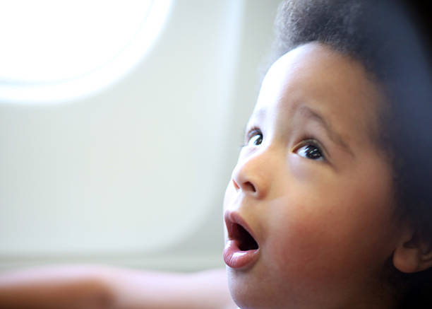 A curious 3-year-old toddler boy sits in a window seat on an airplane, gazing upward with his mouth open in awe and wonder. His wide-eyed expression captures the excitement of flying and the thrill of new experiences through the eyes of a child. A beautiful moment of discovery and innocence at 30,000 feet.